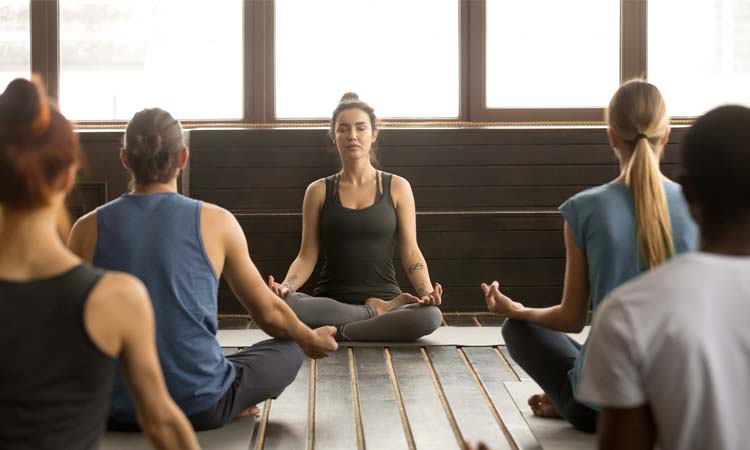 Grupo de personas practicando meditación sentadas en el suelo durante una clase de yoga, con la profesora guiando la práctica en un espacio interior luminoso