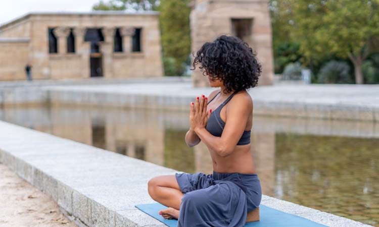 Mujer practicando yoga sentada en postura de meditación con las manos en anjali mudra al aire libre, junto a un estanque, en un entorno tranquilo y natural.