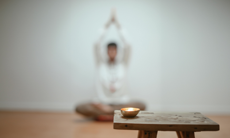 Imagen de un espacio de yoga con una vela encendida en primer plano y una persona sentada en meditación al fondo, creando un ambiente de calma y concentración