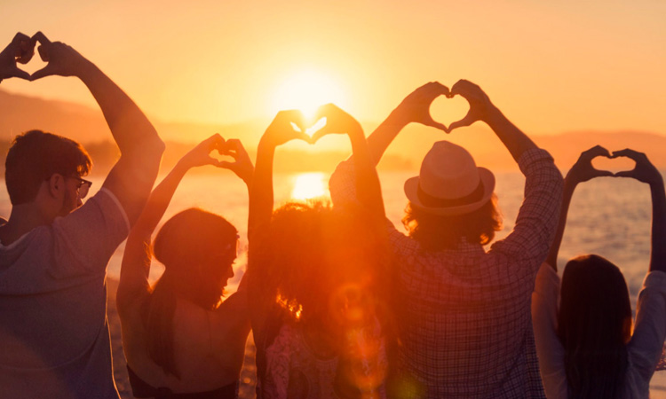 Imagen de un grupo de personas alzando los brazos y formando un corazón con sus manos en un atardecer en la playa representando valores como ahimsa o Bhakti, principios del Jivamukti Yoga